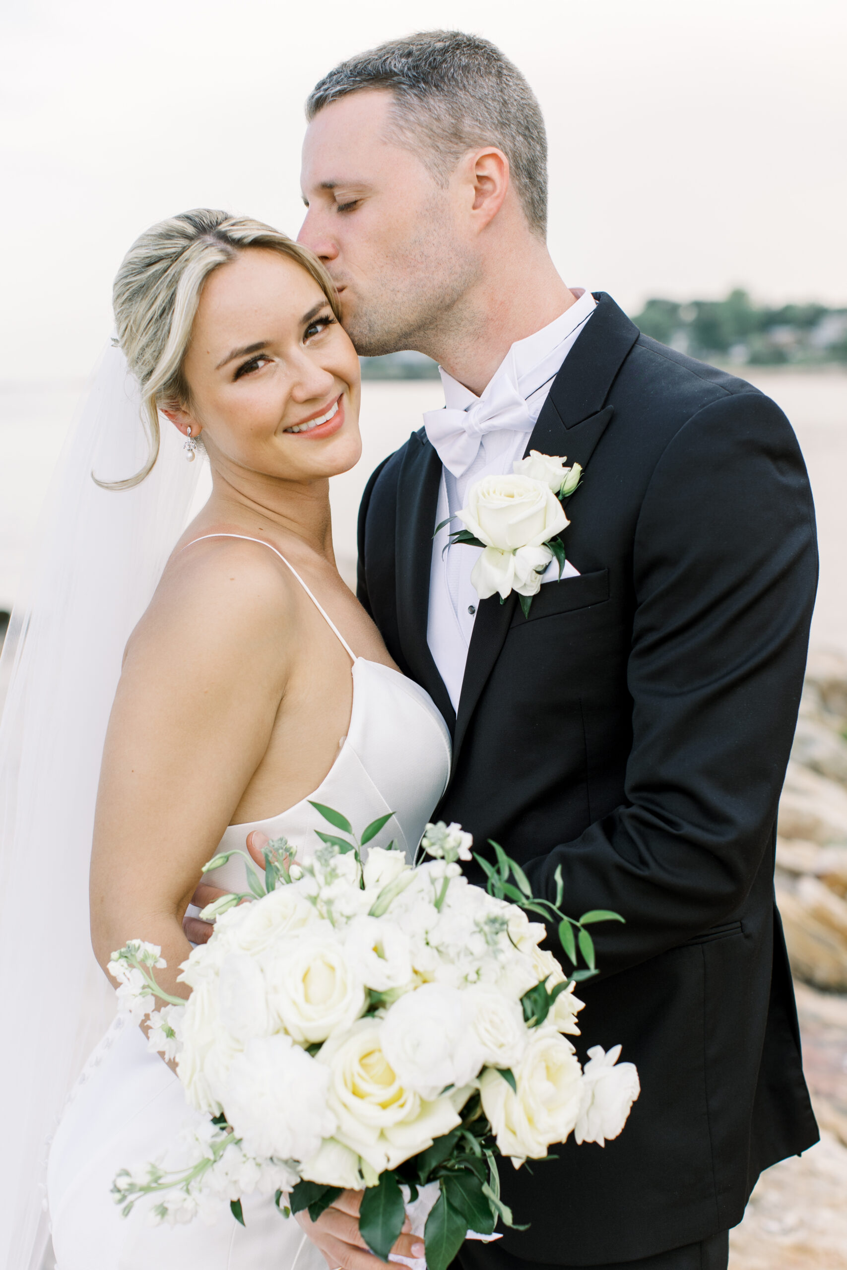 Bride and groom embracing by the waterfront after their Connecticut wedding, featuring romantic bridal makeup and hair styling by A Touch of Color.